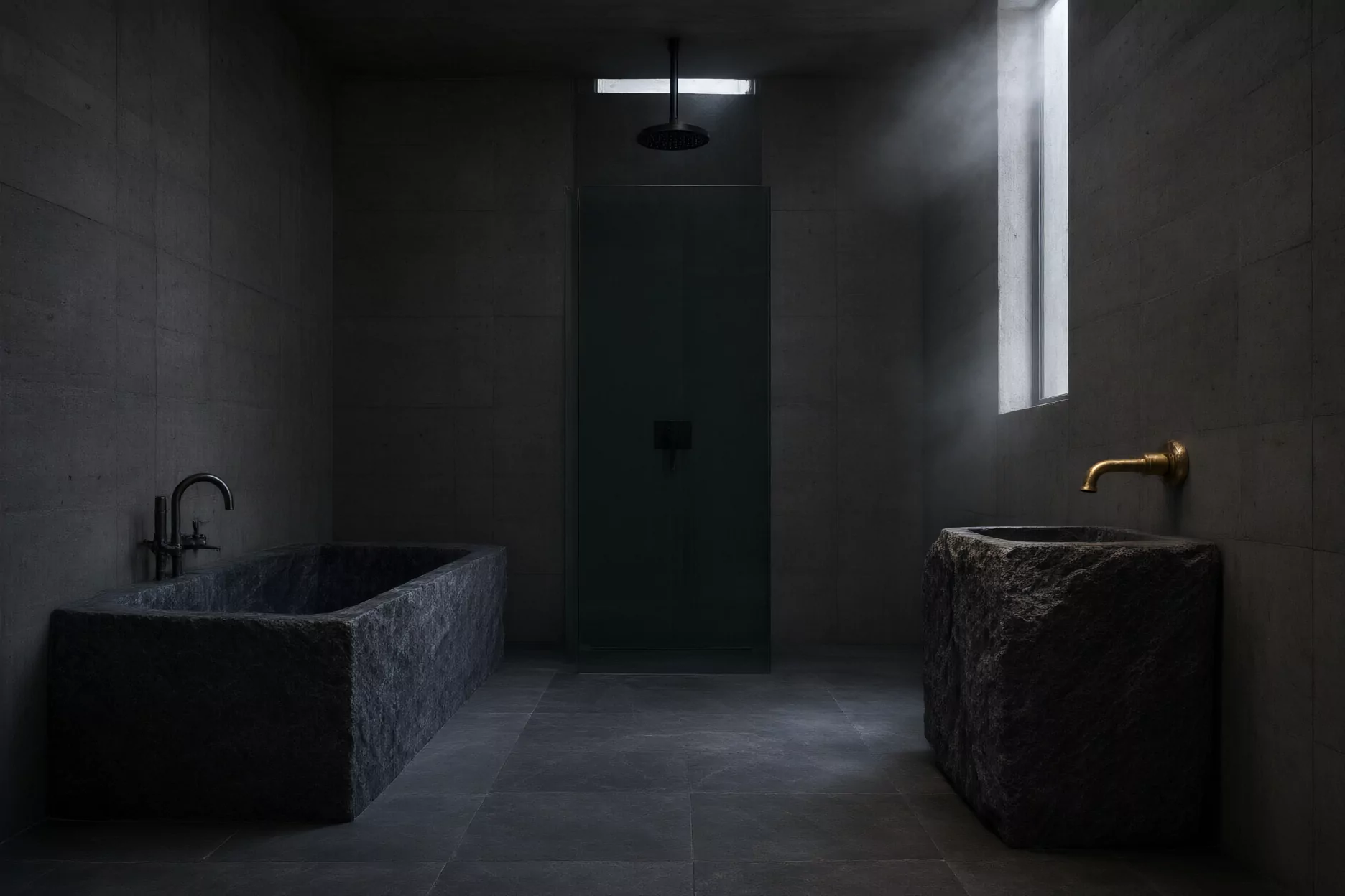 Salle de bain en basalte : baignoire monolithe, douche de plafond, vasque en pierre et robinet laiton.
